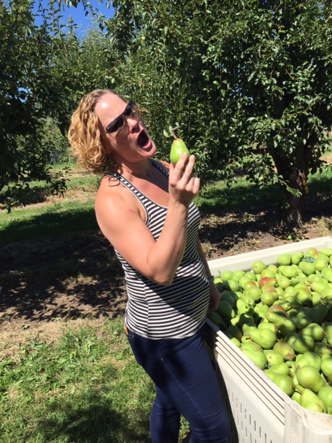 Woman with curly blonde hair in an orchard excitedly about to bite a fresh pear